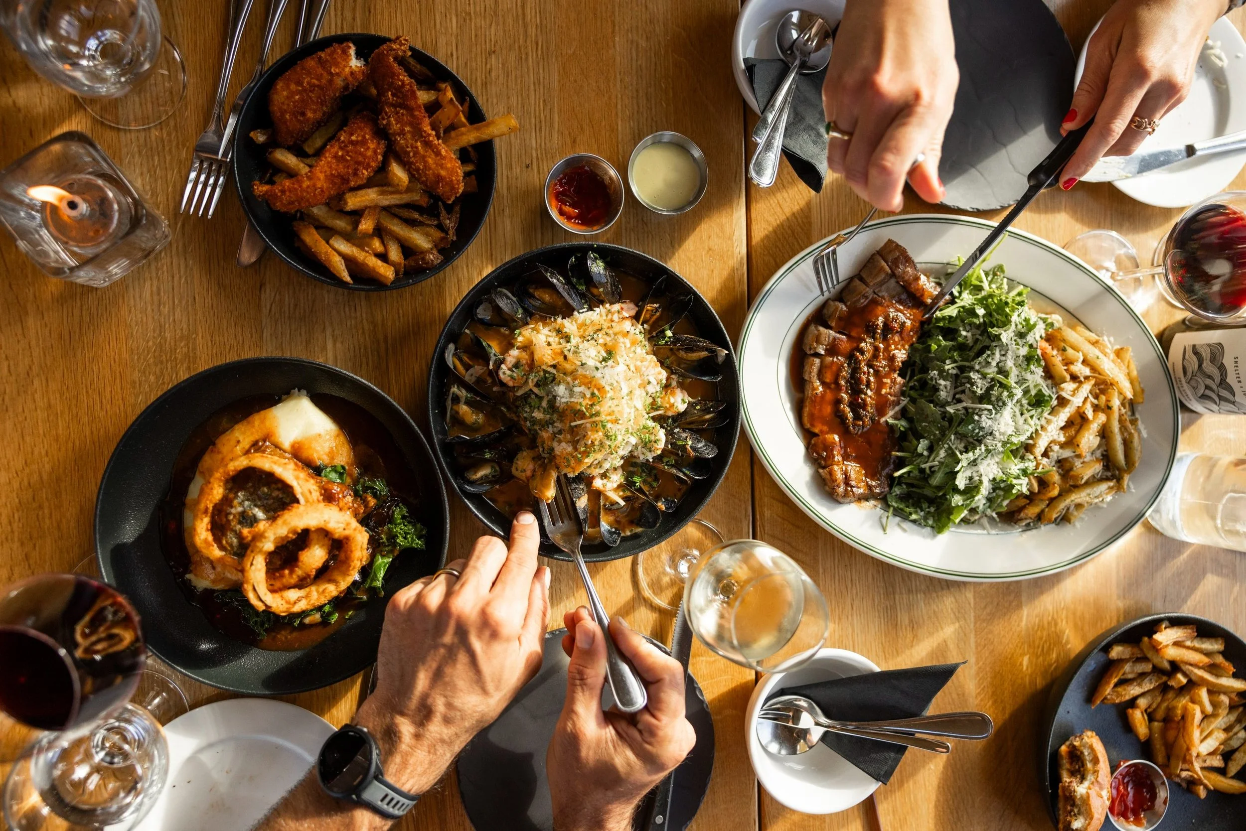 top down view of a table with multiple plates of foodfries fried chicken strips mussels and a steak dishas hands reach in with forks and knives for a shared meal